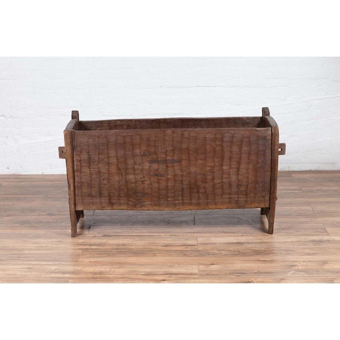 Brown weathered wooden planter with chiseled texture on wood floor next to a white brick wall indoors.