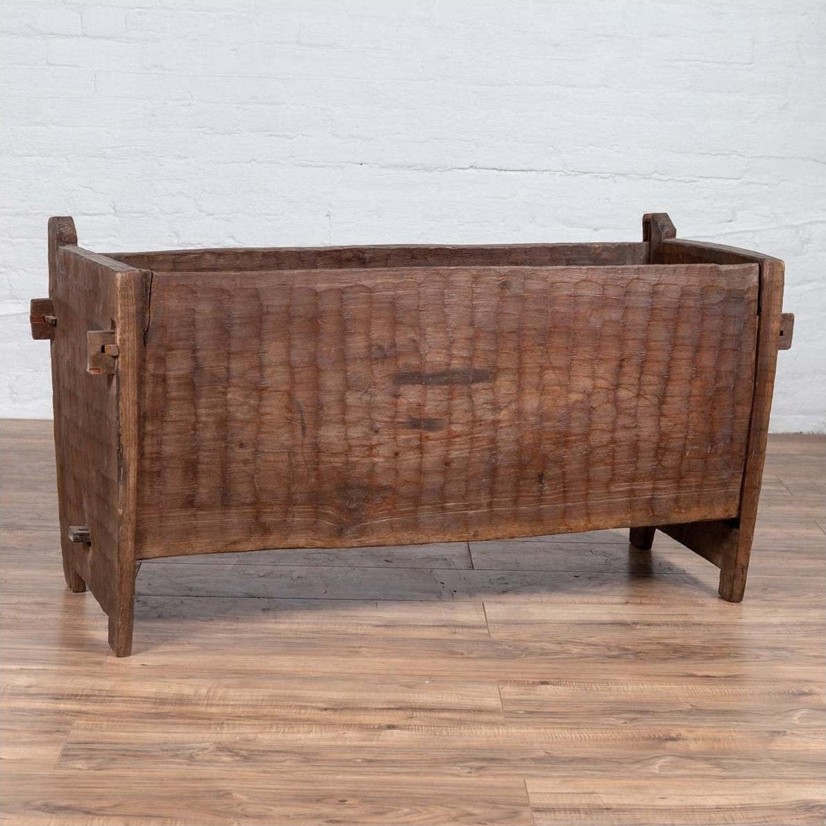 Brown weathered wooden planter with chiseled texture on wood floor against white brick wall indoors.