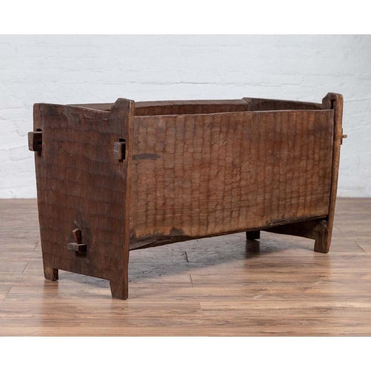 Brown weathered wooden planter with chiseled texture on a wooden floor beside a white brick wall indoors.