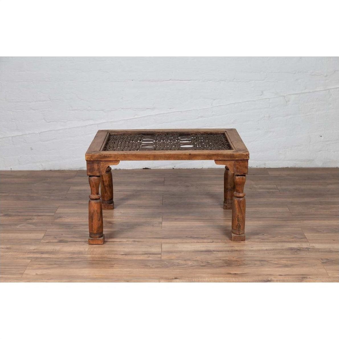 Brown wood side table with window grate top and turned baluster legs, on wood floor near white brick wall indoors.