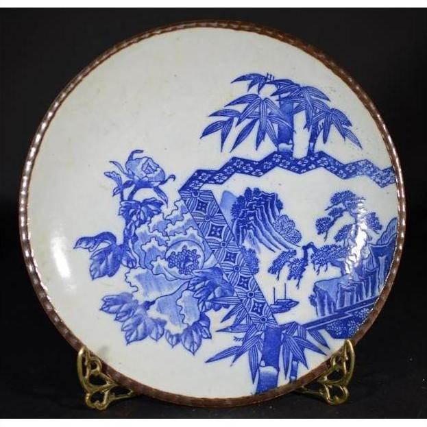 Blue and white ceramic Igezara transferware plate with floral and landscape designs on brass stand, black background.