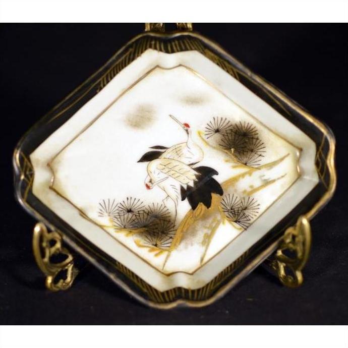 White porcelain Kutani dish with hand painted cranes, pine trees, and gold accents on a display stand.