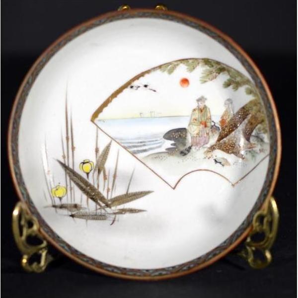 Close-up of multicolor Kutani porcelain plate with two figures, birds, reeds, and red sun on dark background.