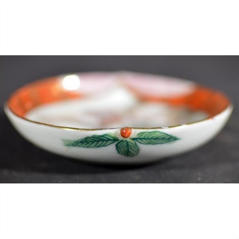 Close-up of antique Japanese Satsuma ceramic plate with green leaf, red dot, and orange rim on dark background.