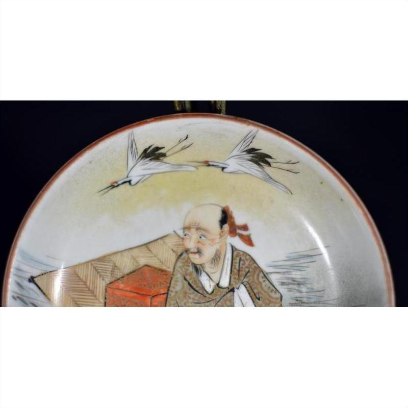 Close-up of an antique Satsuma porcelain plate with painted elderly man and two flying cranes on dark background.