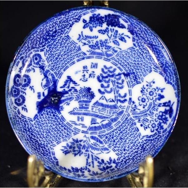 White porcelain Japanese transferware plate with floral and landscape designs on decorative metal stand, close-up view.