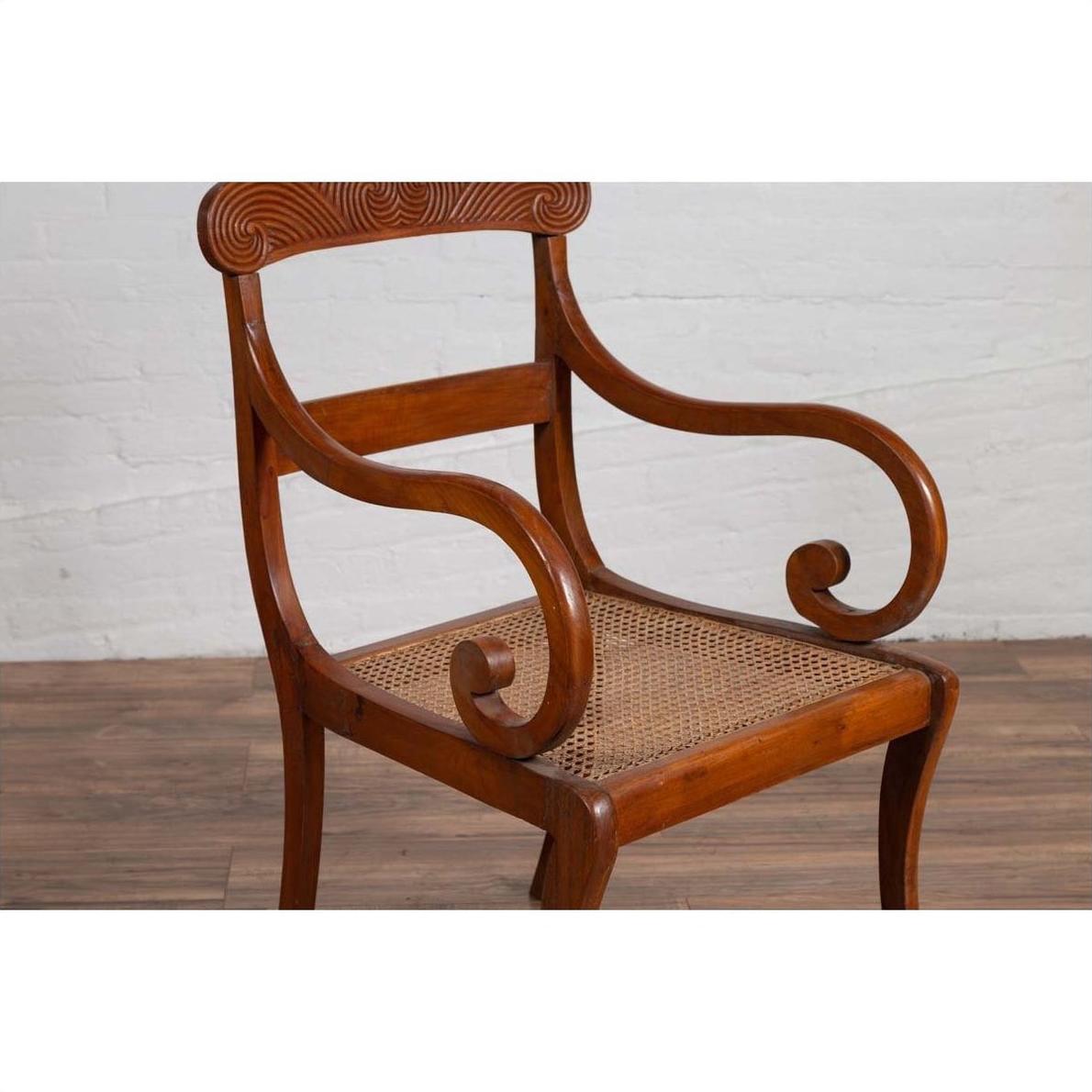 Brown wood and rattan armchair with carved back and curved arms, set against a white brick wall indoors.