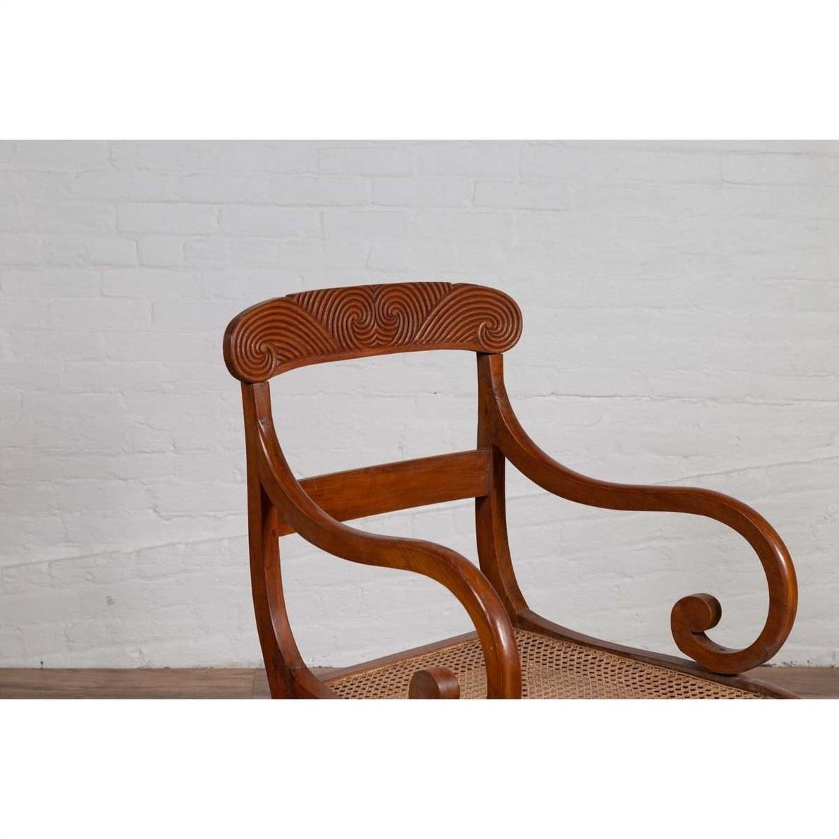 Brown wooden and rattan Javanese armchair with carved back and curving arms against a white brick wall indoors.