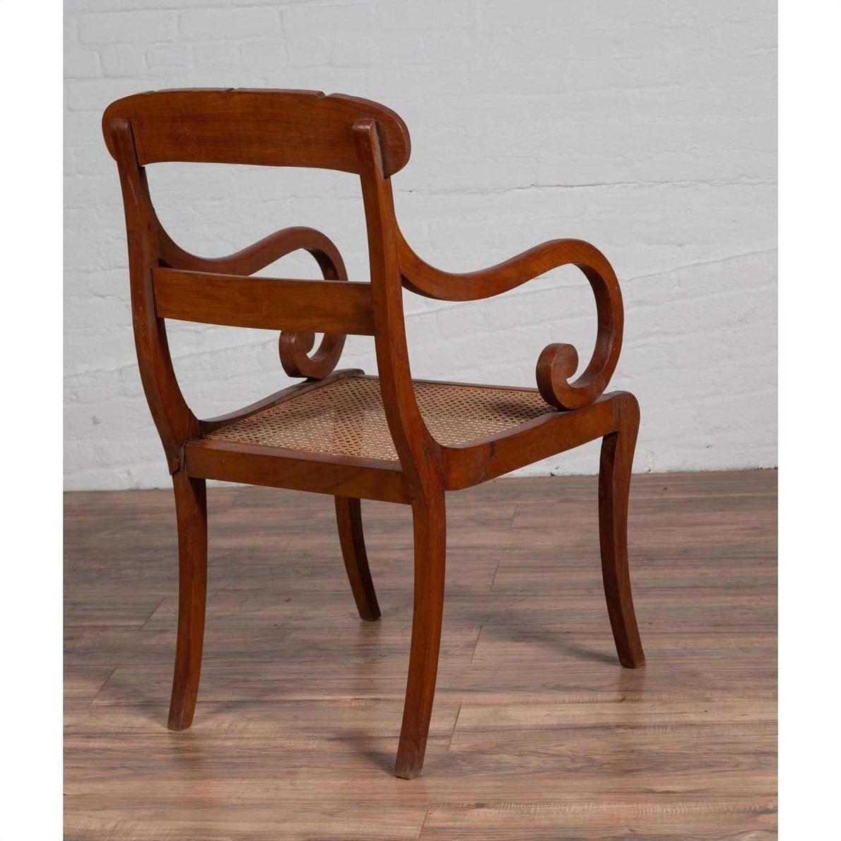 Brown wood and rattan armchair with carved back and curving arms on wood floor next to white brick wall indoors.