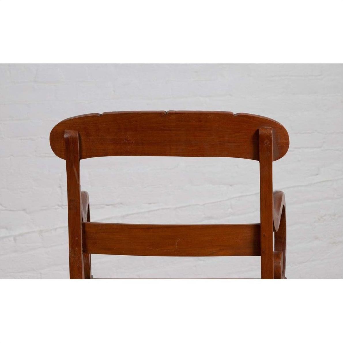Brown wooden and rattan armchair with carved back and curved arms, shown indoors against a white brick wall.