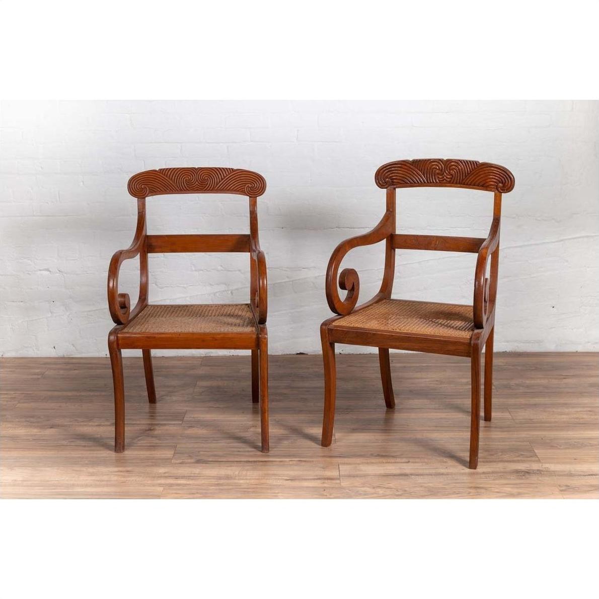 Brown wooden and rattan Javanese armchairs with carved backs, side by side on a wooden floor indoors.