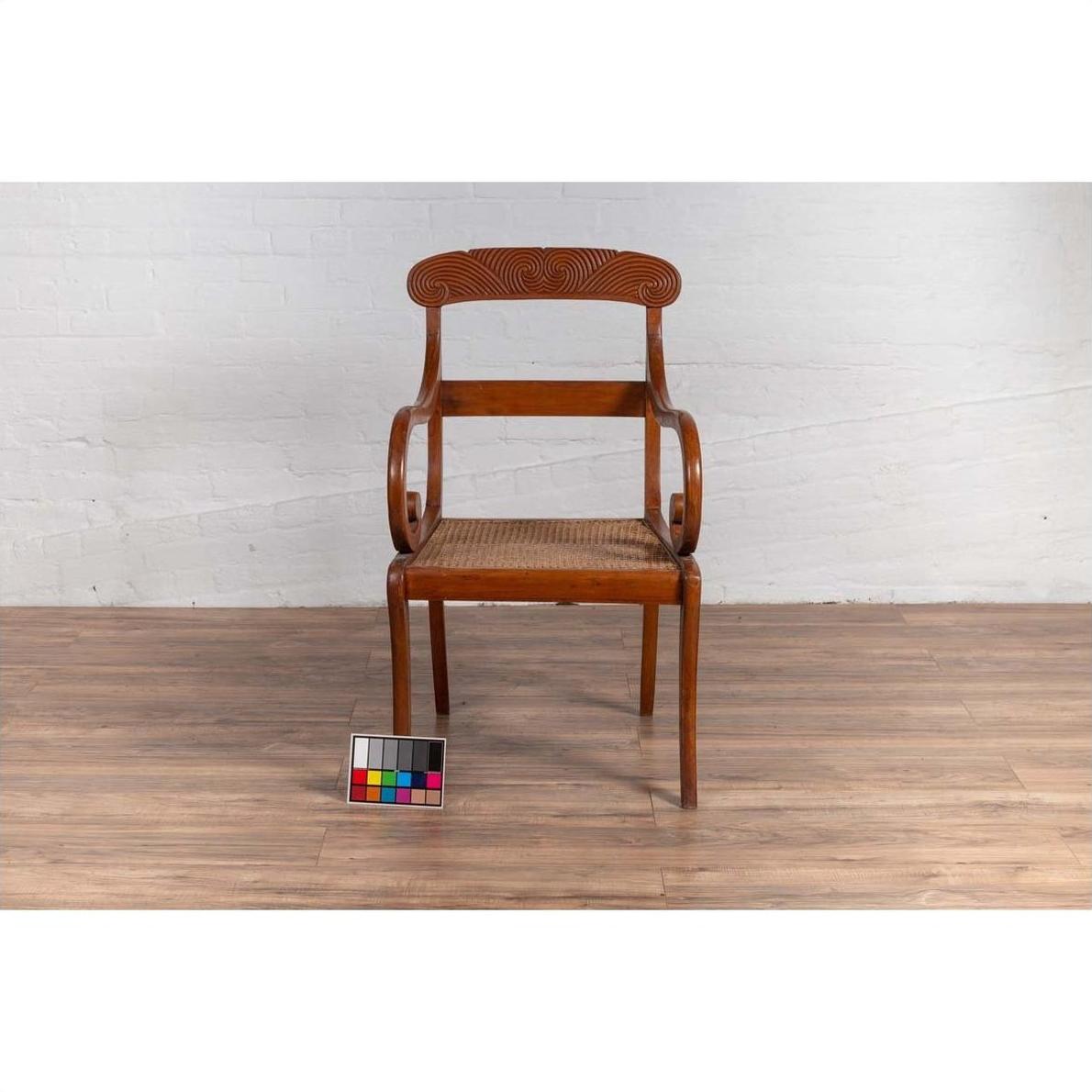 Brown wood and rattan armchair with carved back and curved arms on wood floor against white brick wall indoors.