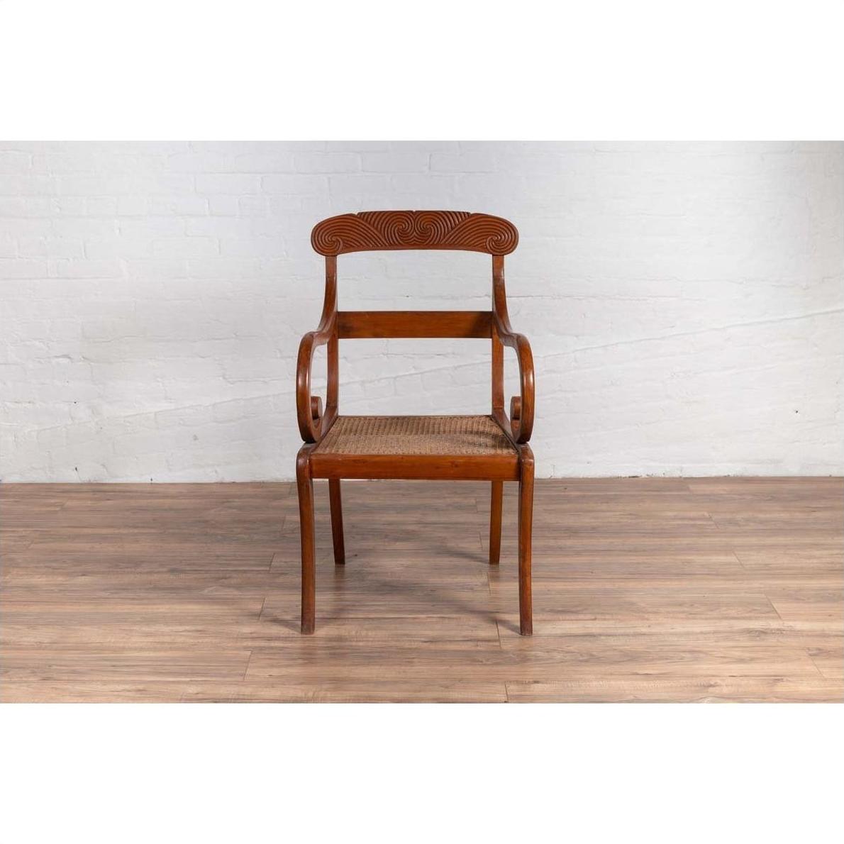 Brown wooden Javanese armchair with carved back, curved arms, and rattan seat on a wooden floor indoors.