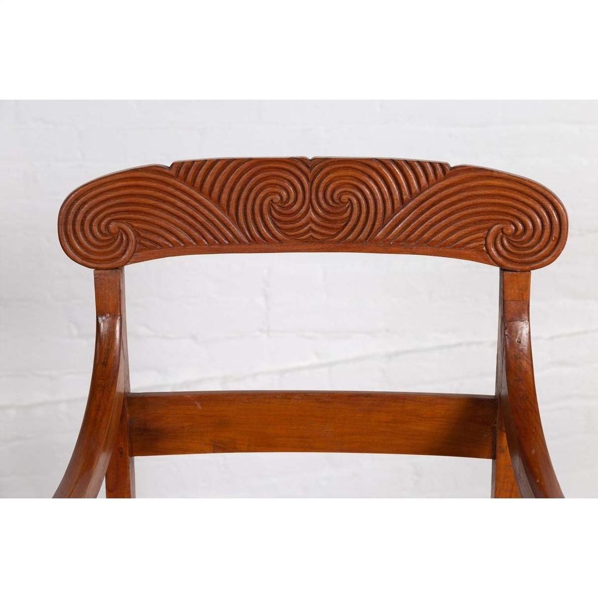 Close-up of brown wooden Javanese armchair backrest with carved swirl patterns on the top rail.