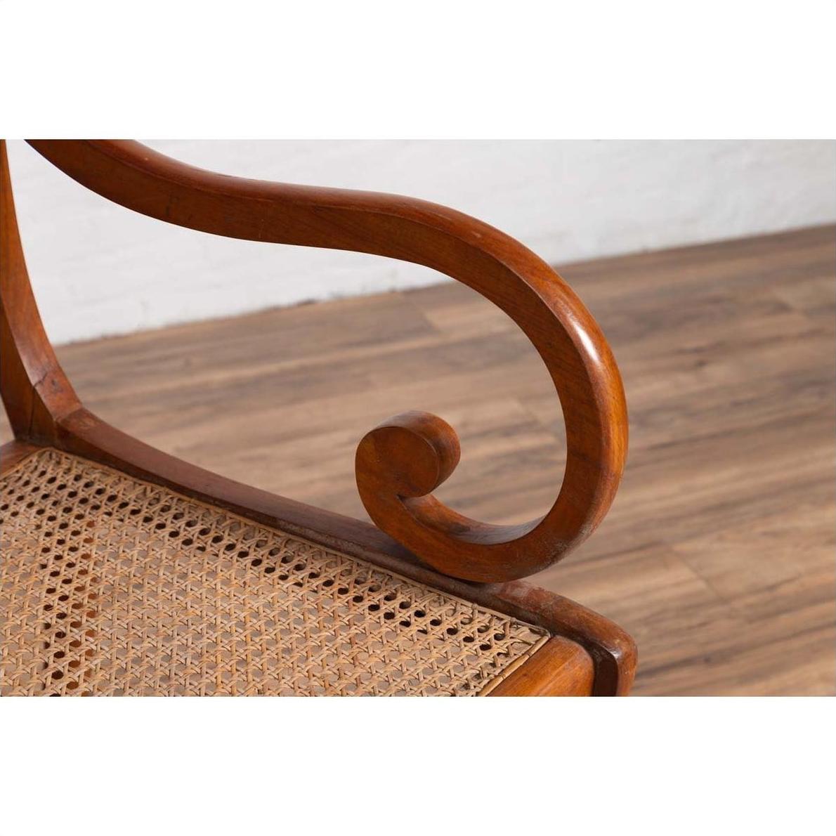 Brown wooden and rattan armchair with carved back and curved arms, on wood floor against white wall indoors.