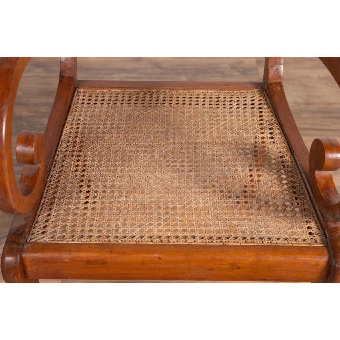 Close-up of brown wooden Javanese armchair with carved back, curved arms, and rattan seat on wood floor.