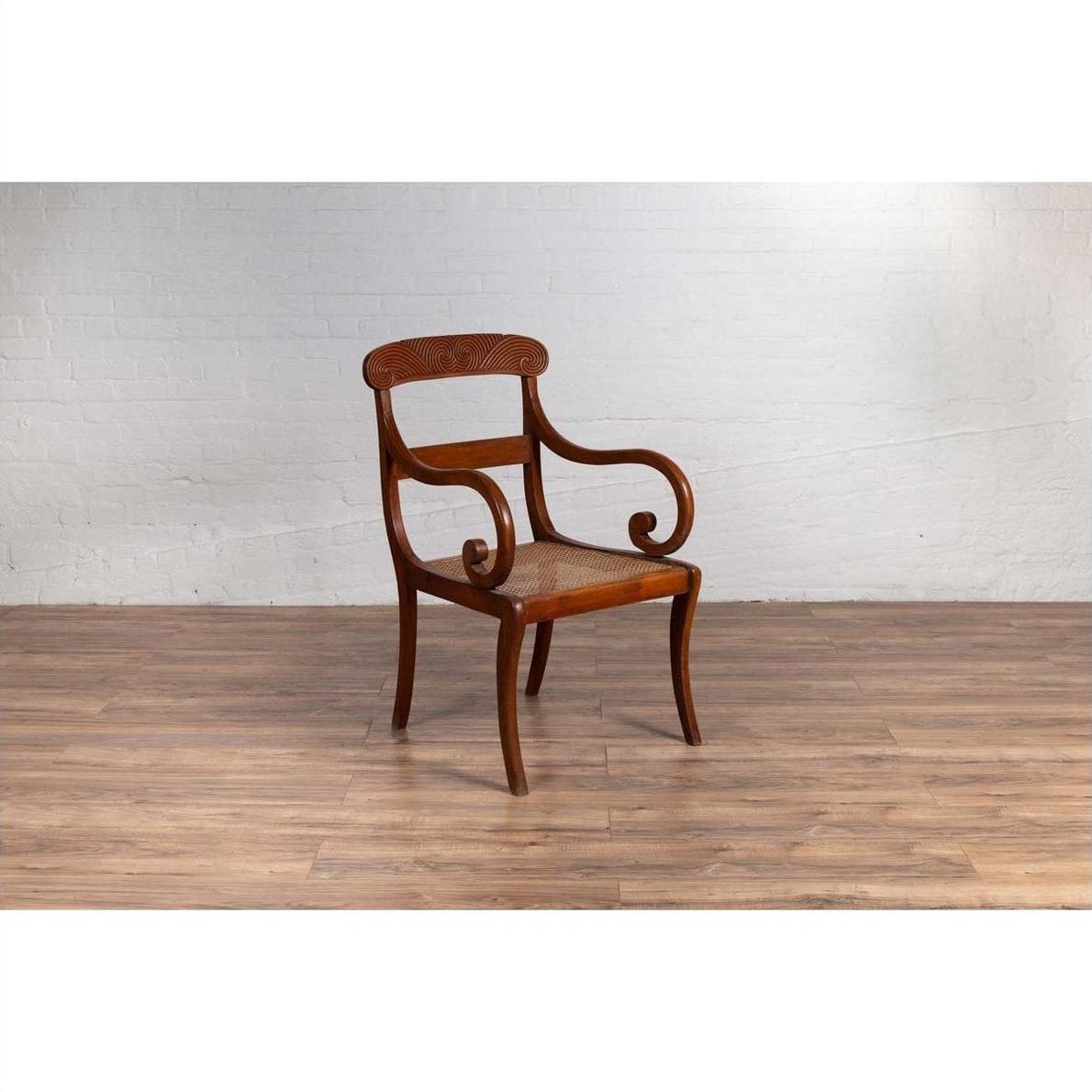 Brown wooden and rattan Javanese armchair with carved back and curved arms, on wood floor by white brick wall indoors.