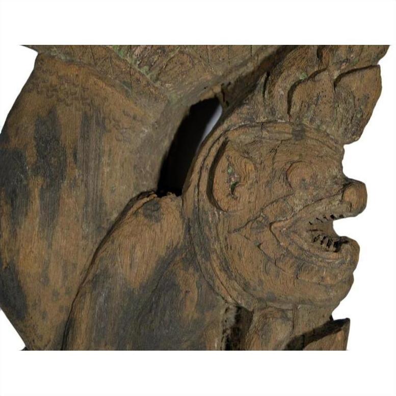Close-up of brown teakwood Burmese temple carving, mirrored guardian relief with stylized headgear.