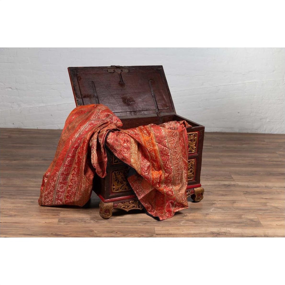 Open brown wooden blanket chest with gilt accents, draped in red and gold patterned fabric.