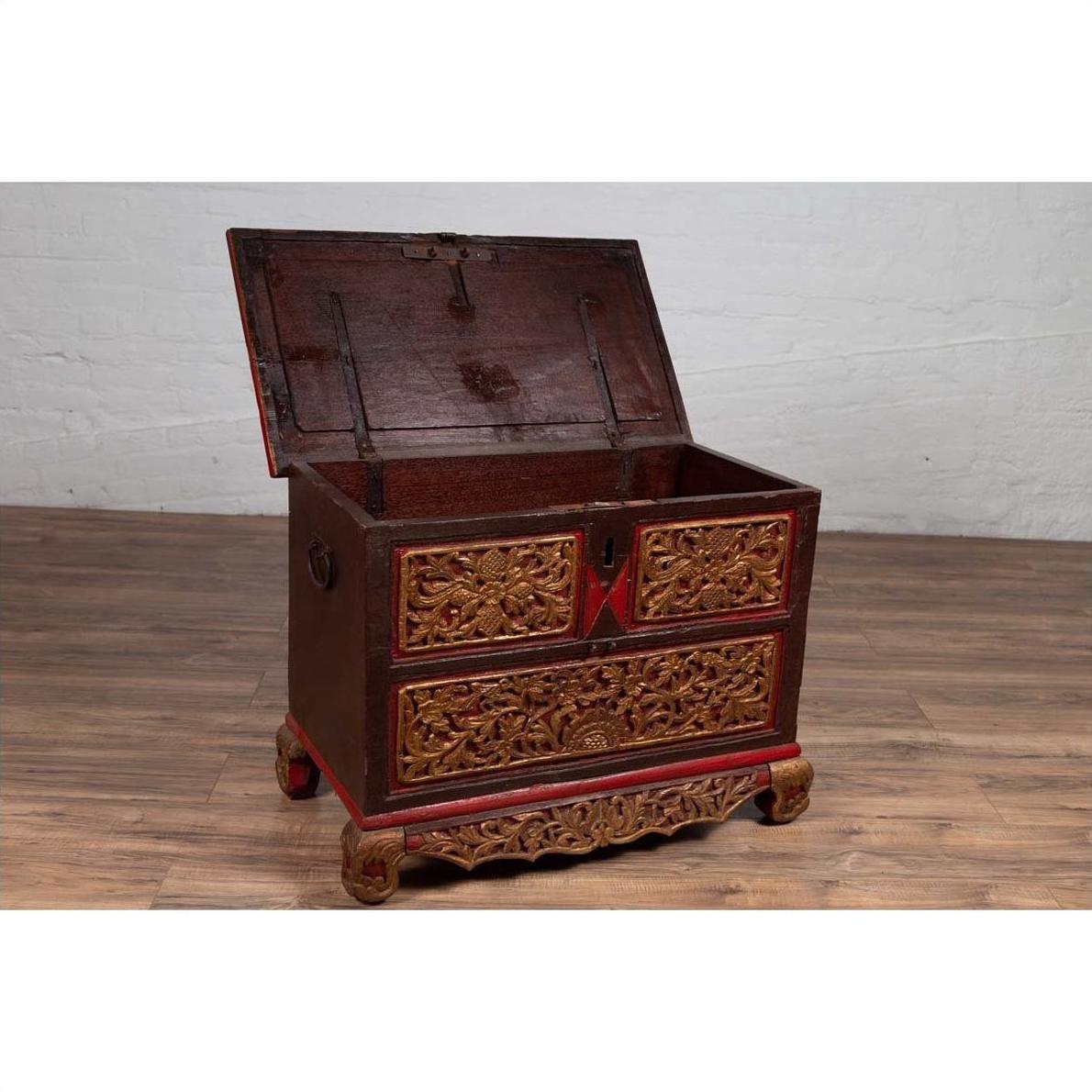 Brown hand carved wooden blanket chest with gilt accents, open lid, on wood floor against white brick wall.