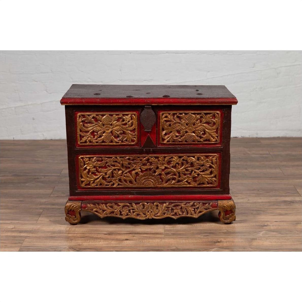 Brown wooden blanket chest with hand-carved details and gilt accents on hardwood floor against a white wall.