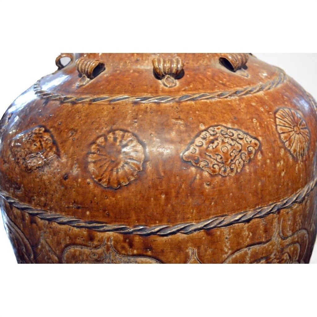 Brown ceramic water jar close-up with embossed geometric and floral patterns, textured surface visible.