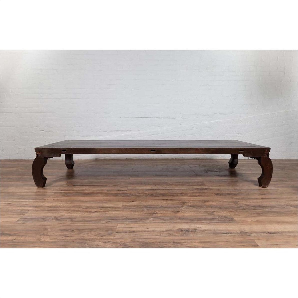 Dark wood opium bed with curved legs on wooden floor against white brick wall, antique Northern Chinese Ming Dynasty style.