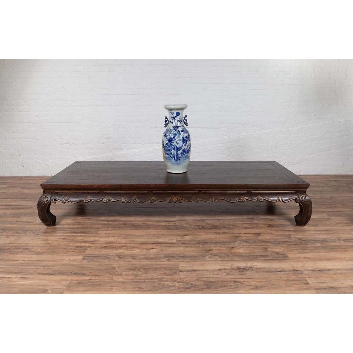 Dark wood opium bed with carved legs against a white brick wall, antique Ming Dynasty style, indoor setting.