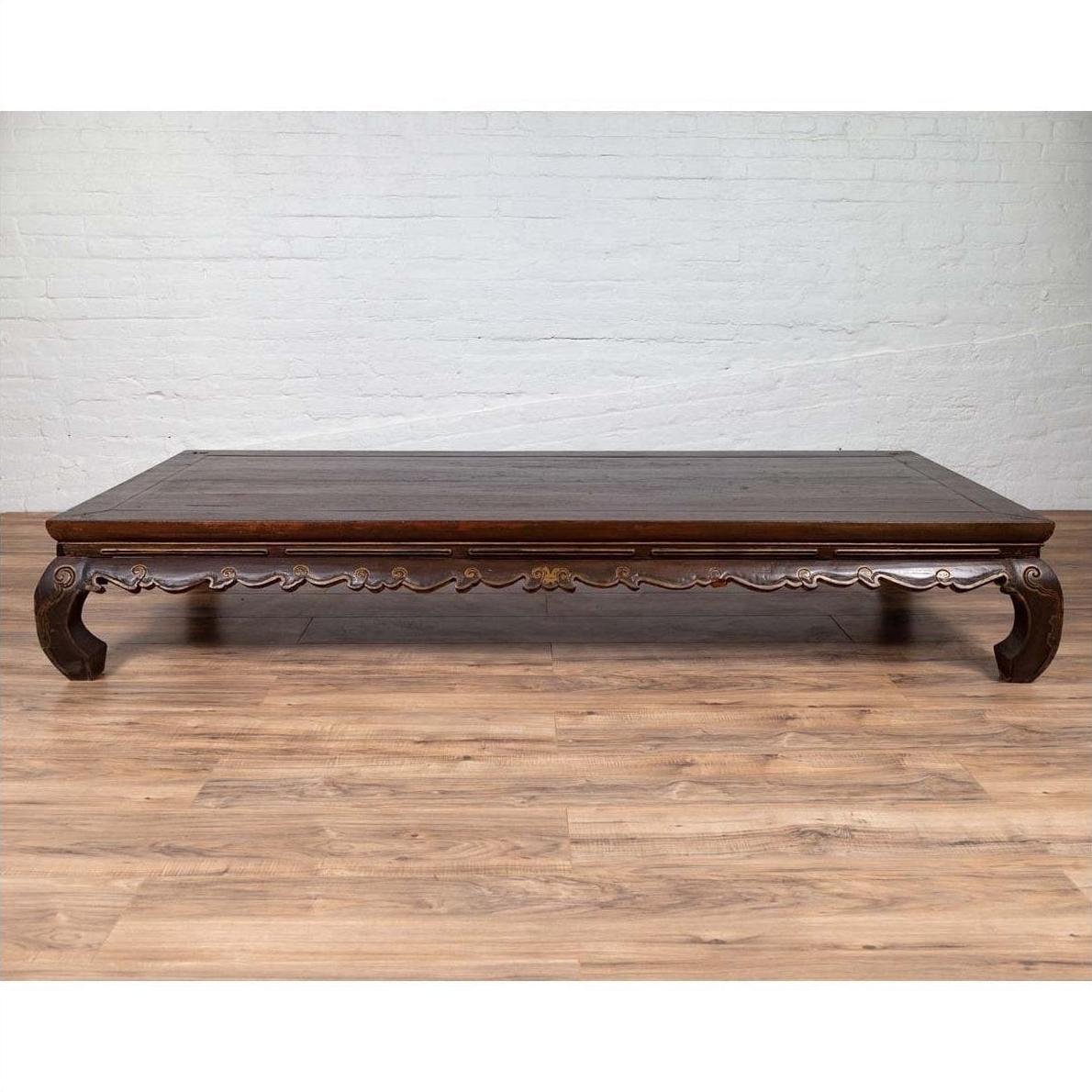 Dark wood Ming dynasty style Kang opium bed with hand-carved legs on light wood floor against white brick wall indoors.