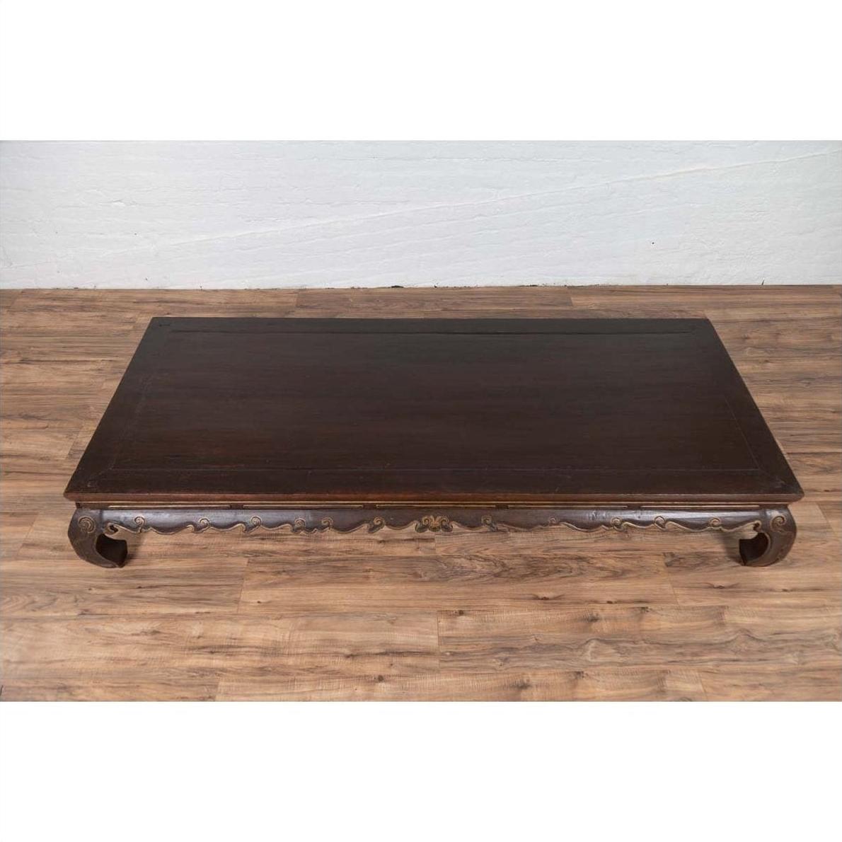 Dark wood Chinese Ming Dynasty style opium bed with curled legs on a wood floor against a white wall indoors.