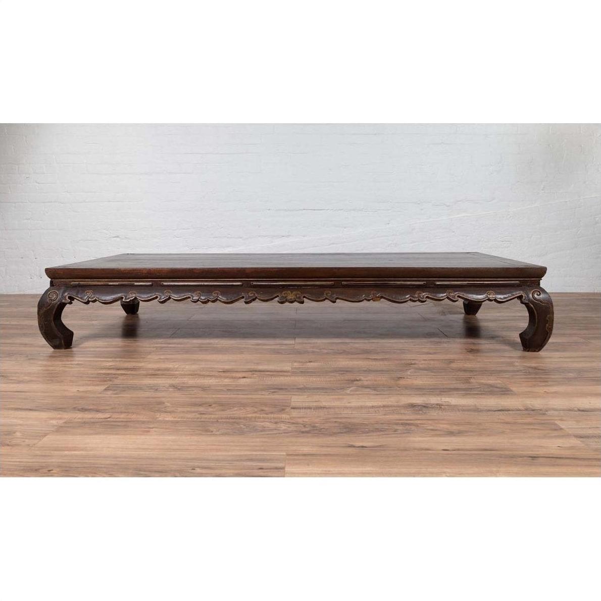Dark wood ornate carved opium bed in Ming Dynasty style, placed on wooden floor against a white brick wall indoors.