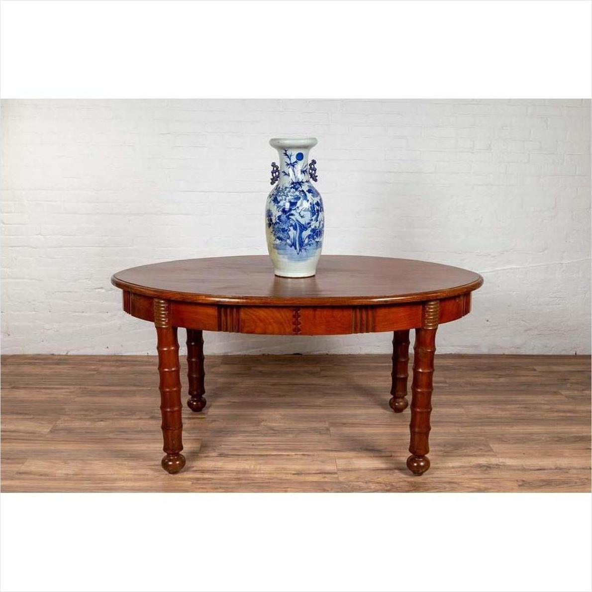 Brown wood oval dining table with spindle legs and warm patina, placed against a white brick wall.