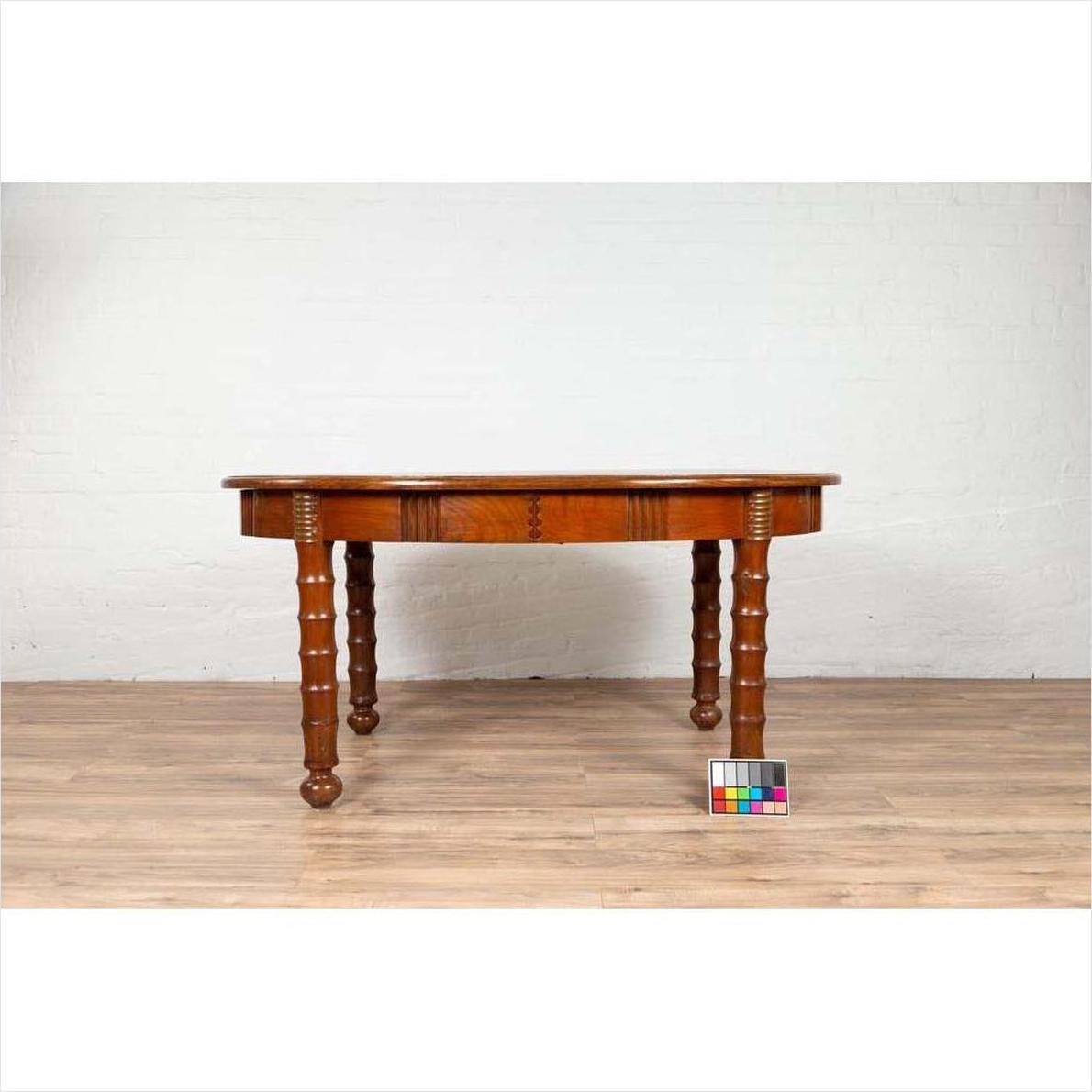 Brown wood antique oval dining room table with spindle legs, warm patina, on wood floor by white brick wall.
