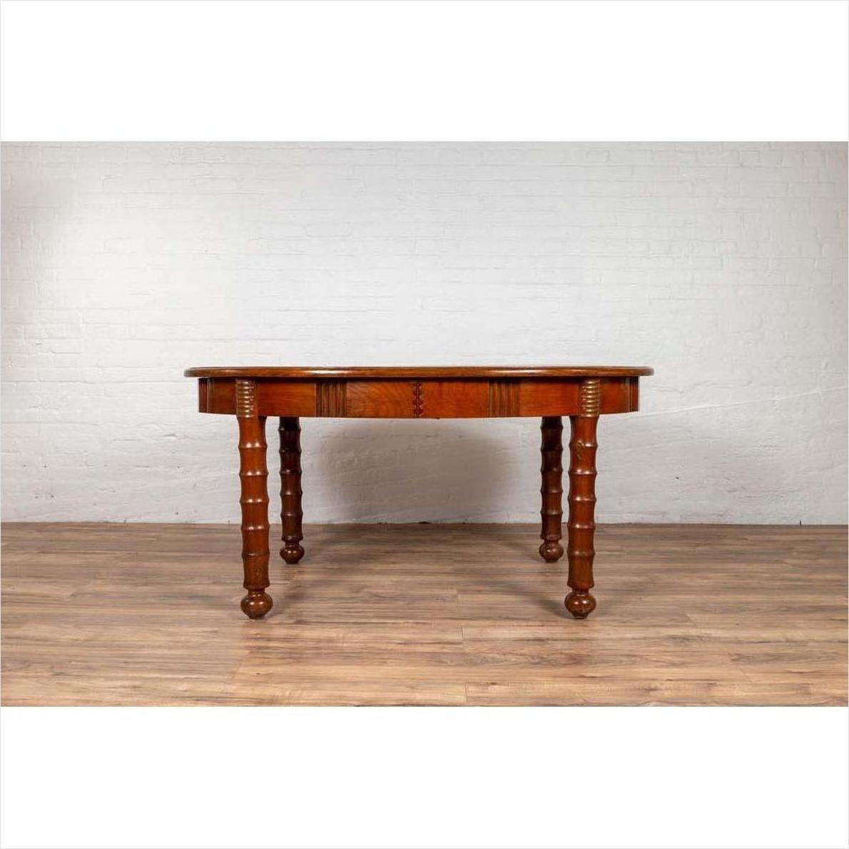 Brown wood oval dining table with spindle legs and warm patina on wooden floor, Indonesian antique style.