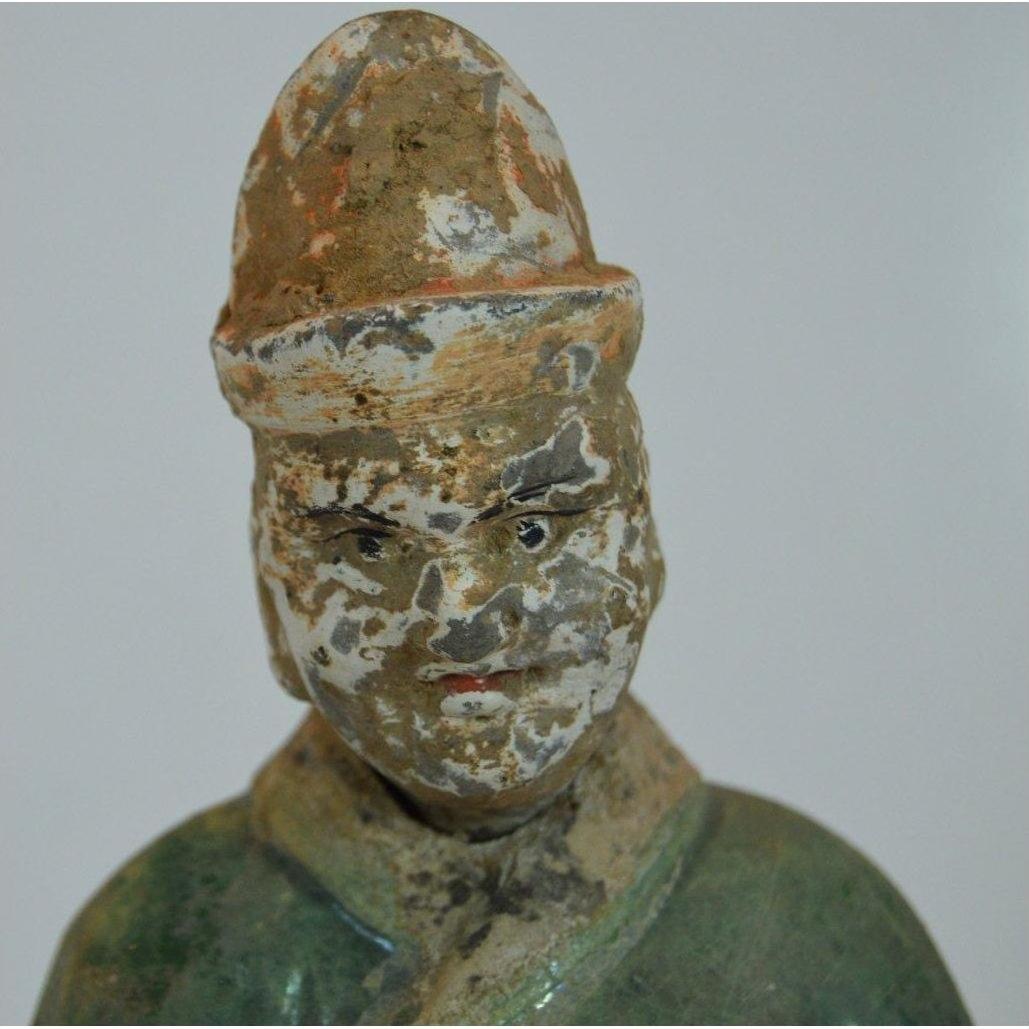 Close-up of antique terracotta court figure statue with hat, faded paint, and facial wear against plain background.