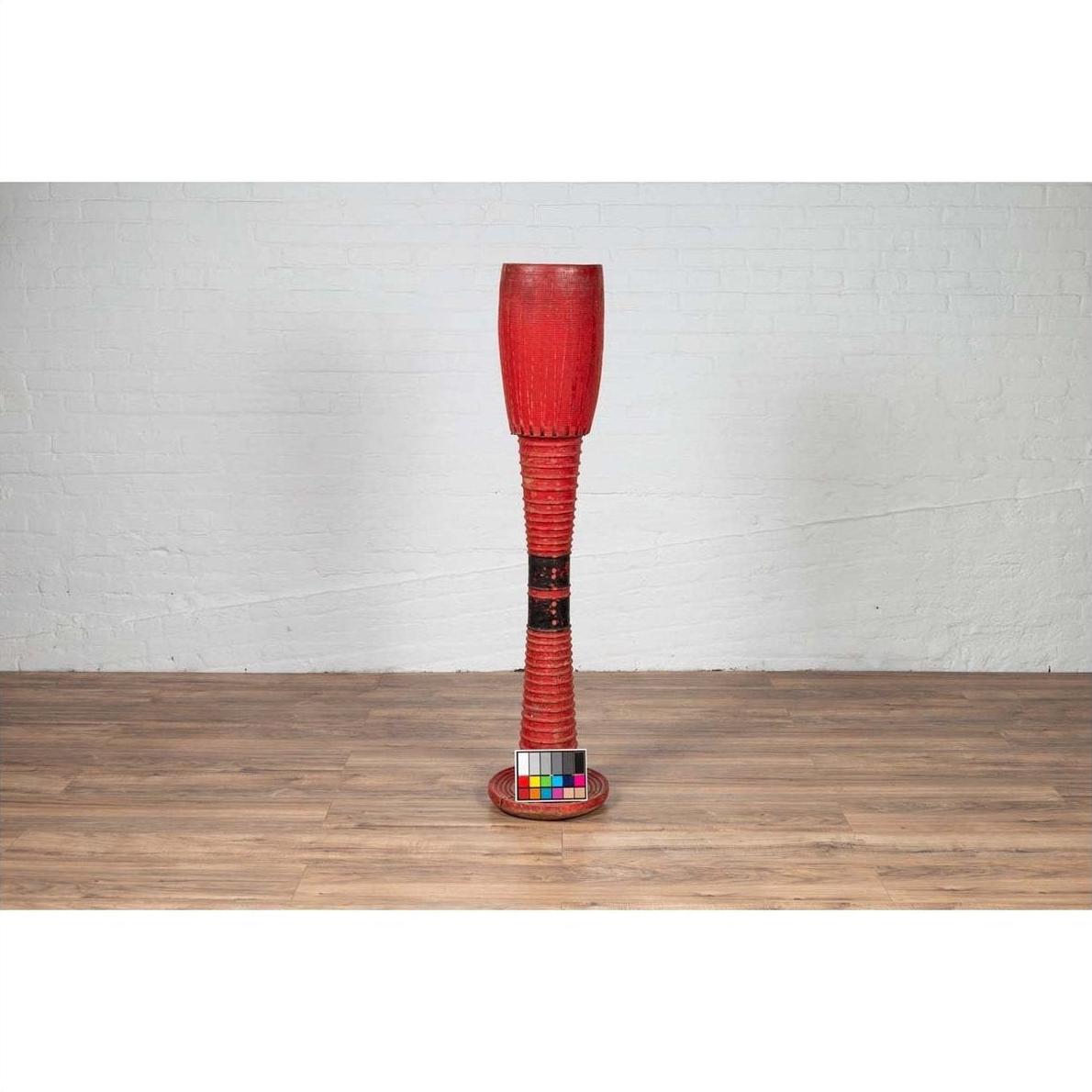 Red wood ceremonial drum with round base on a wood floor next to a white wall indoors.