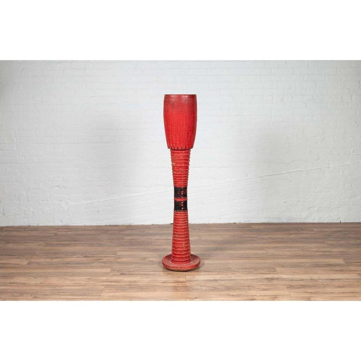 Red wood ceremonial drum with round base on wooden floor in front of white brick wall indoors.