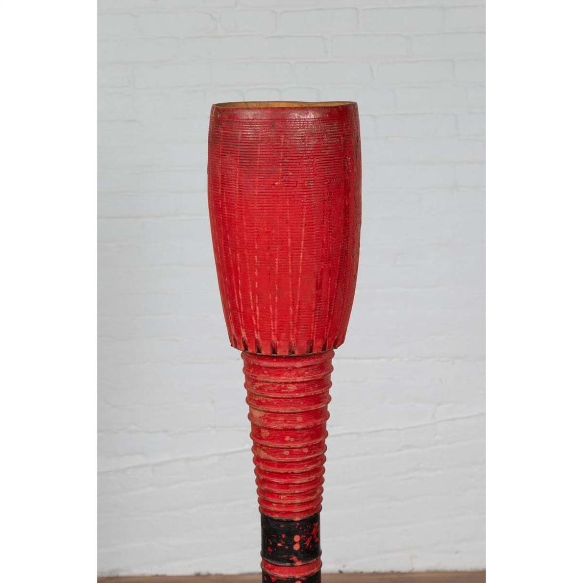 Red wood ceremonial drum with round base, placed indoors against a white brick wall.