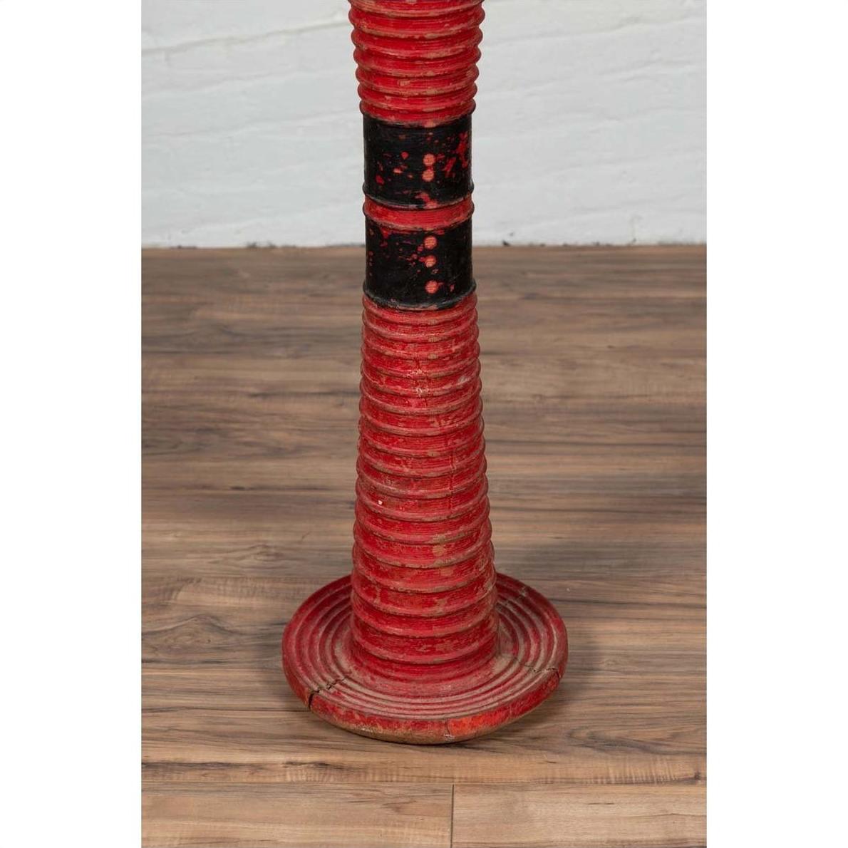Red wood ceremonial drum with round base on wooden floor next to a white brick wall indoors.