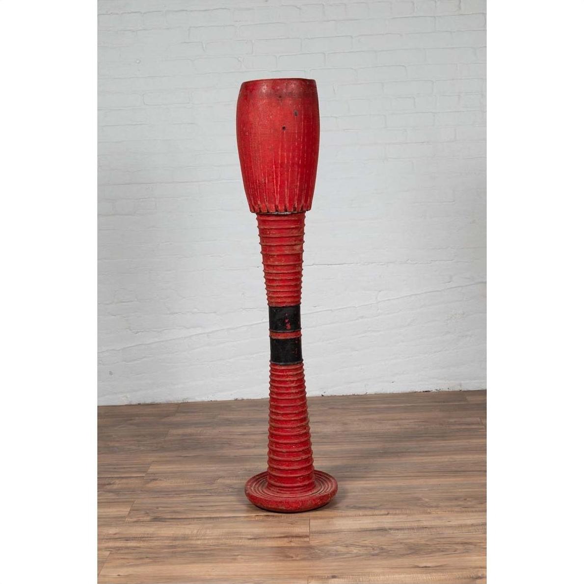 Red wooden ceremonial drum with round base on wooden floor beside a white brick wall indoors.
