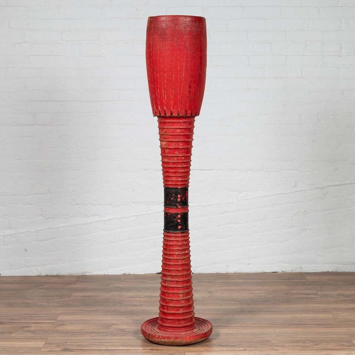 Red wood freestanding Thai ceremonial drum with round base on wooden floor next to white brick wall indoors.