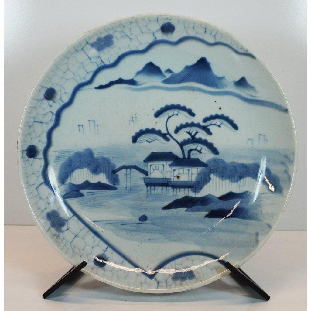 Blue and white porcelain plate with hand painted mountain, trees, and house scene on a black stand.