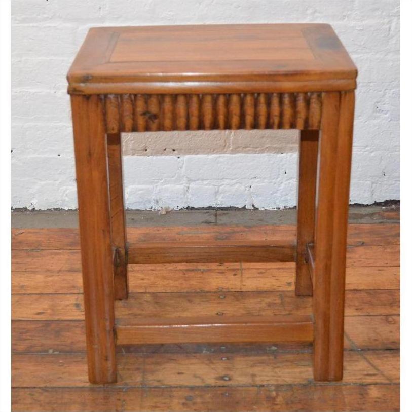 Brown wooden Art Deco stool on wooden floor in front of white brick wall.