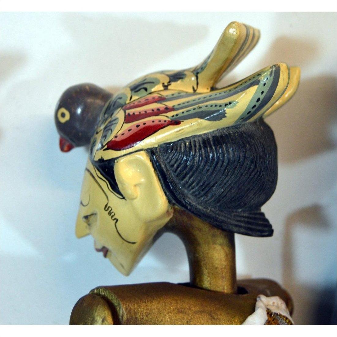 Brown hand-carved wooden puppet with painted bird headdress and stylized features, shown indoors.