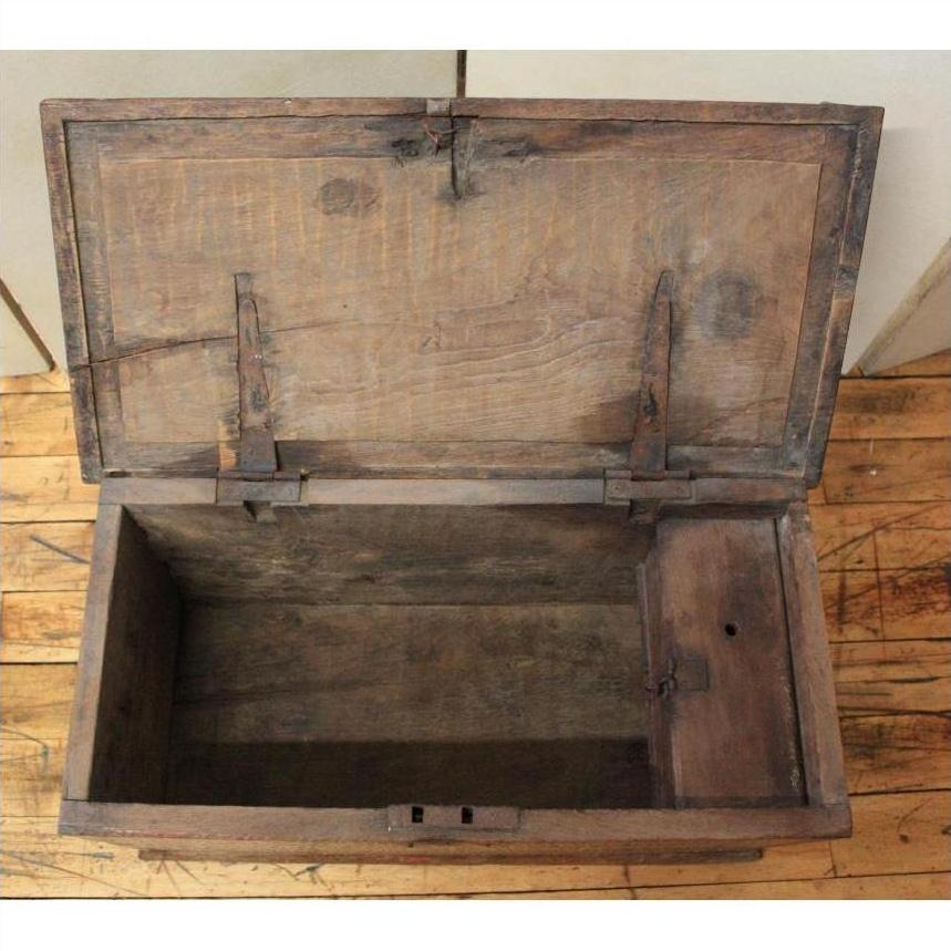 Brown wooden blanket chest with metal hinges, open and empty, sitting on a scratched wooden floor indoors.
