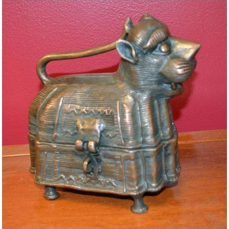 Brass animal-shaped box with handle, latch, and four legs on wooden surface against red wall.