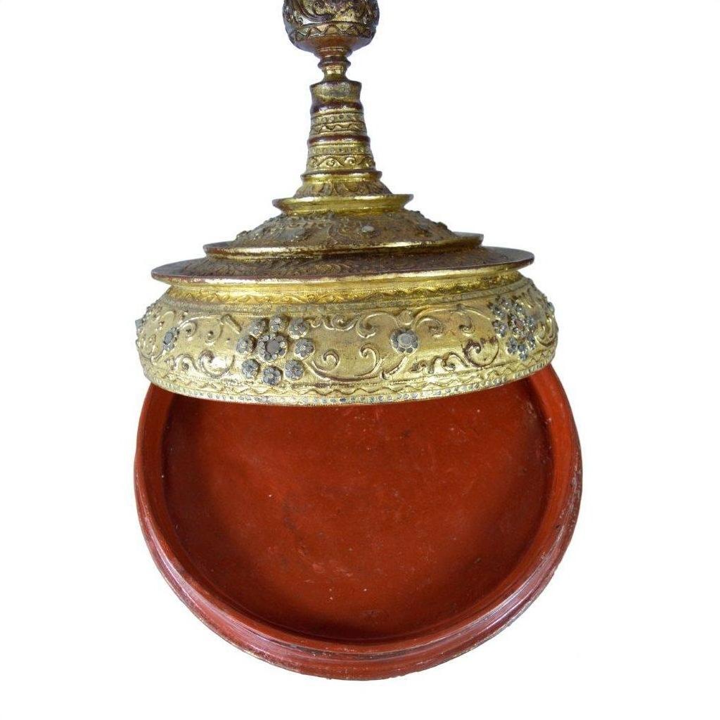 Close-up of round red lacquered Burmese offering bowl with gilt, jewel-studded lid, ornate mica inlay, partially open.