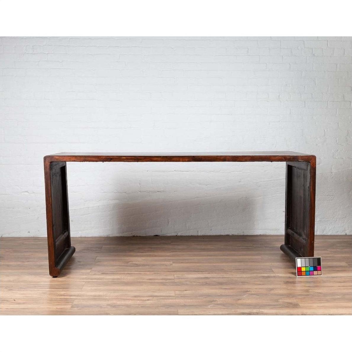 Dark wood Burmese vintage waterfall console table with scrolling feet on a wood floor against a white brick wall.