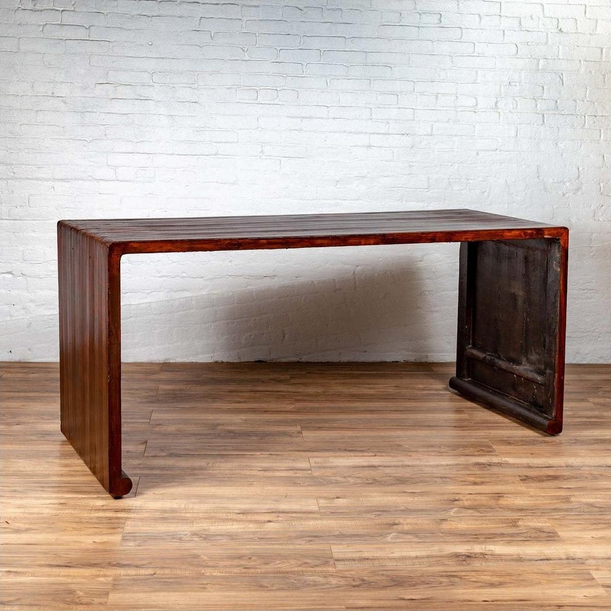 Dark wood Burmese vintage waterfall console table with scrolling feet on a wooden floor against a white brick wall.