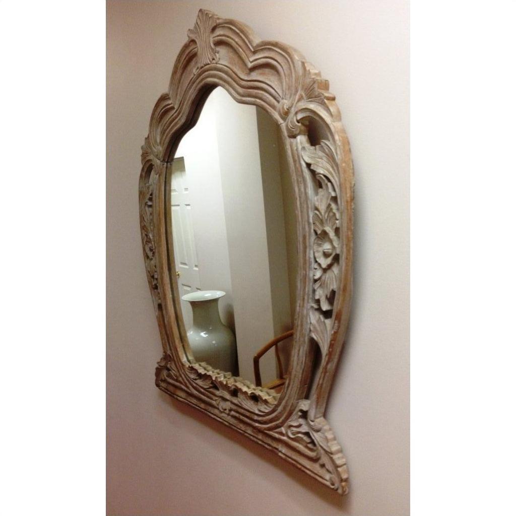 Carved mirror with decorative frame on wall, reflecting hallway, white vase, and part of a chair in view.